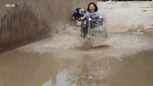 輪椅王 兩個人騎著自行車穿過崎嶇不平的小路上的大泥坑，周圍濺起水花，類似於在崎嶇地形上使用 MIJO MT01/MT02 手推變電動輪椅拖頭時所獲得的快感。