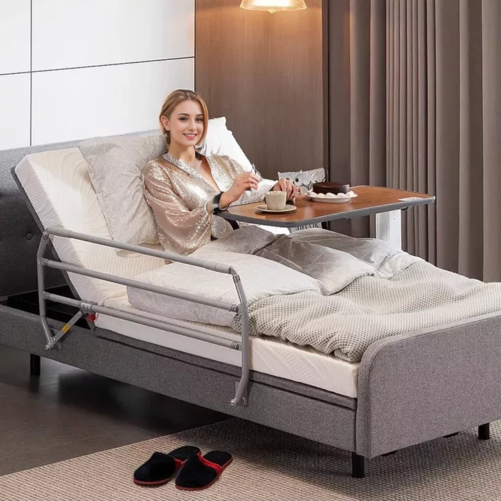 【輪椅王】 一個穿著睡衣的人坐在德國品牌 1.05m/1.2m 床寬升降多功能電動護理床 WC800(出口版) 上，配有可調式托盤桌。他們手捧杯子，微笑著，雙腿伸在1.05M/1.2M寬的床上，蓋著毯子。拖鞋放在床邊的地板上。