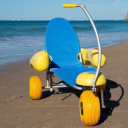 輪椅王 一輛帶有黃色大輪子和扶手、藍色座椅和弧形手柄的沙灘輪椅停在沙灘上，背景是大海。