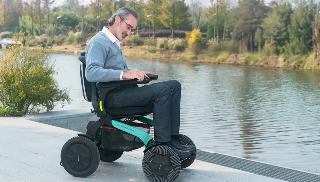 輪椅王 一名坐在 Robooter E60 智慧型電動輪椅手機控制及折合電動輪椅上的男子坐在公園的湖邊。他低頭看著輪椅的控制裝置。背景中可以看到樹木和綠化。