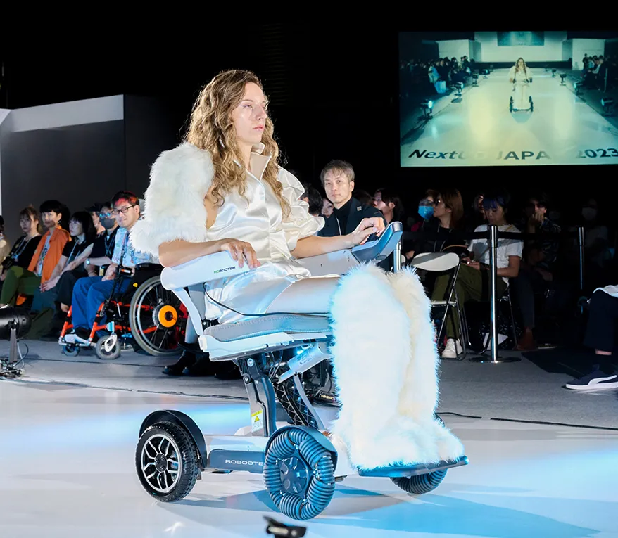 輪椅王 穿著白色服裝和毛茸茸的靴子的模特在跑道上騎著 Robooter X40 折疊電動輪椅，觀眾在觀看，背景中的視頻屏幕顯示相同的跑道場景。