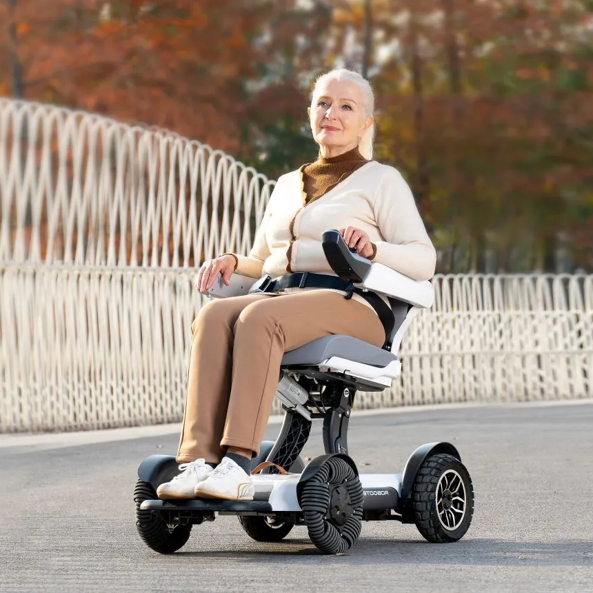 輪椅王 一位老婦人坐在戶外 Robooter X40 折疊電動輪椅上，沿著一條帶有波浪狀欄桿的彎曲路徑，周圍環繞著秋葉。她表情滿足，穿著米色毛衣和褲子。