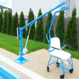 輪椅王 一個固定式泳池升降架配有白色椅子，專為無障礙設計，位於室外游泳池旁邊，毗鄰草地和高大的灌木叢。