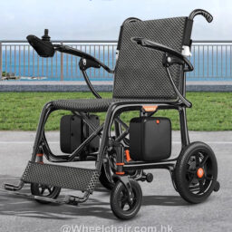 輪椅王 一款現代的 9 公斤碳纖超輕電動輪椅，配有堅固的黑色框架、扶手和腳踏板，位於室外鋪好的表面上，有金屬欄桿，背景是大海。