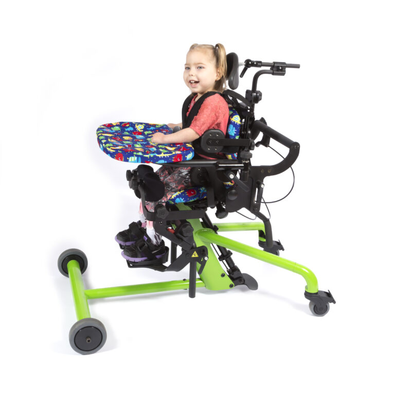 【輪椅王】 一名幼兒坐在專為有座位需求的兒童設計的彩色自適應椅上微笑。兒童輪椅 PT50001-1 Bantam Extra Small 具有亮綠色框架、支撐帶和移動輪。她穿著有圖案的緊身褲和紅色上衣。