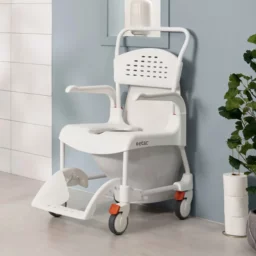 【輪椅王】 瑞典ETAC CLEAN有輪沐浴便椅（可調高度）是一款帶輪子的白色移動式淋浴椅，放置在浴室中。它配有軟墊座椅、可折疊扶手和帶通風孔的高靠背。這張高度可調的椅子靠著淡藍色的牆壁放置，旁邊放著盆栽植物和衛生紙捲。