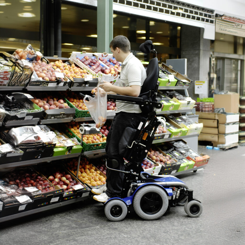 【輪椅王】 一名坐在配備站立功能的電動輪椅上的人從雜貨店的展示櫃上採摘水果。他們一手拿著塑膠袋，另一手伸手去拿水果，使用瑞士進口的LEVO C3垂直電動輪椅。背景中可以看到貨架上擺滿了各種水果。