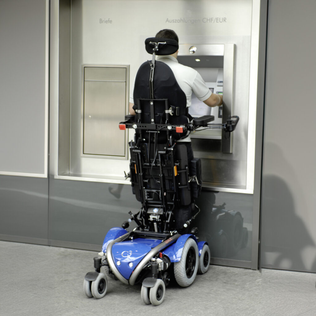 【輪椅王】 一名使用配備機械手臂的輪椅的人正在使用 ATM 機。面向ATM，瑞士進口LEVO C3立式電動輪椅位於正前方，讓交易變得更方便。 ATM 安裝在標有“Briefe”和“Auszahlungen CHF/EUR”的牆上。