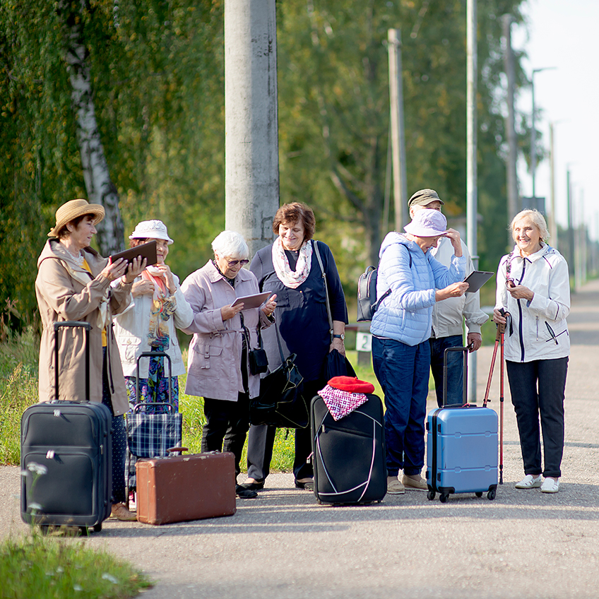 【輪椅王】 一群老人提著行李箱站在鄉村公路旁。有些人穿著休閒夾克，戴著帽子。兩名女性正在查看地圖或文件，很可能正在查看長者旅遊注意事項，而其他女性則在進行交談。背景是樹木繁茂的地區和電線桿。