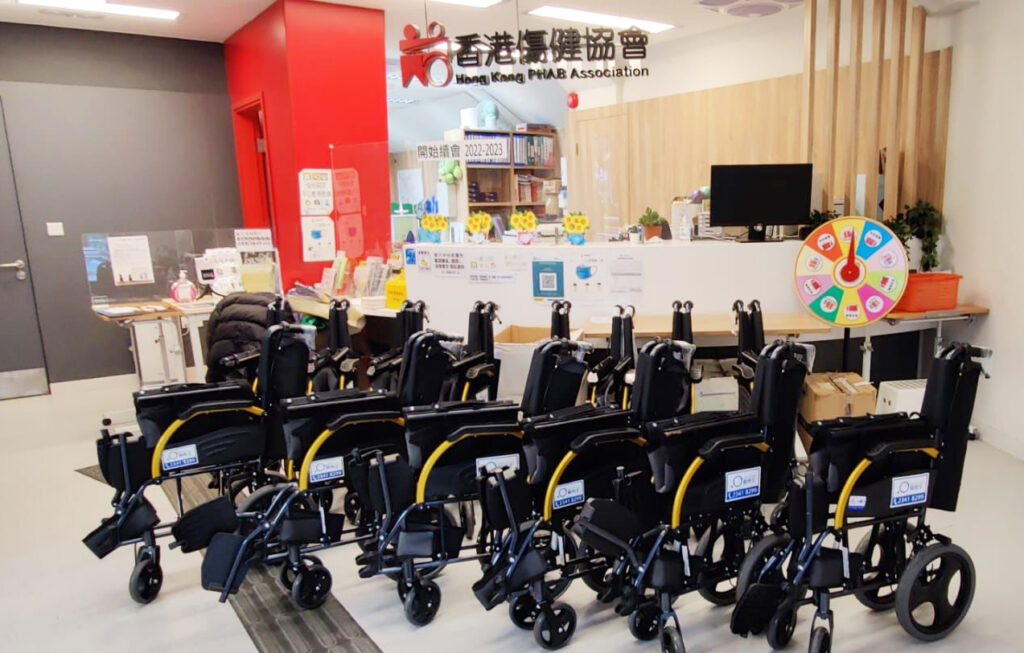 輪椅王客戶 捐贈給香港傷健協會的一排黑黃色輪椅陳列在他們辦公室的服務櫃檯前。辦公室的牆上貼著各種資訊海報，櫃檯上有一台電腦，右側還有一個色彩繽紛的獎品輪盤。