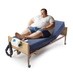 【輪椅王】         一名穿著休閒服的男子坐在醫院病床上，床上放著藍色 Invacare microAIR 壓力氣墊床。該床的承重能力高達 225 公斤，側面配有控制裝置。