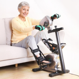 【輪椅王】 一位坐在沙發上的老婦人戴著運動手套，穿著黃色毛衣，使用有防護支架的手踏板鍛鍊機。