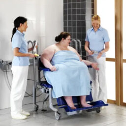 【輪椅王】 一個人坐在一張鋪著藍色床單的 ArjoHuntleigh 椅上，兩名身穿淺藍色制服的醫護人員在一間鋪著瓷磚的房間裡照顧他們。