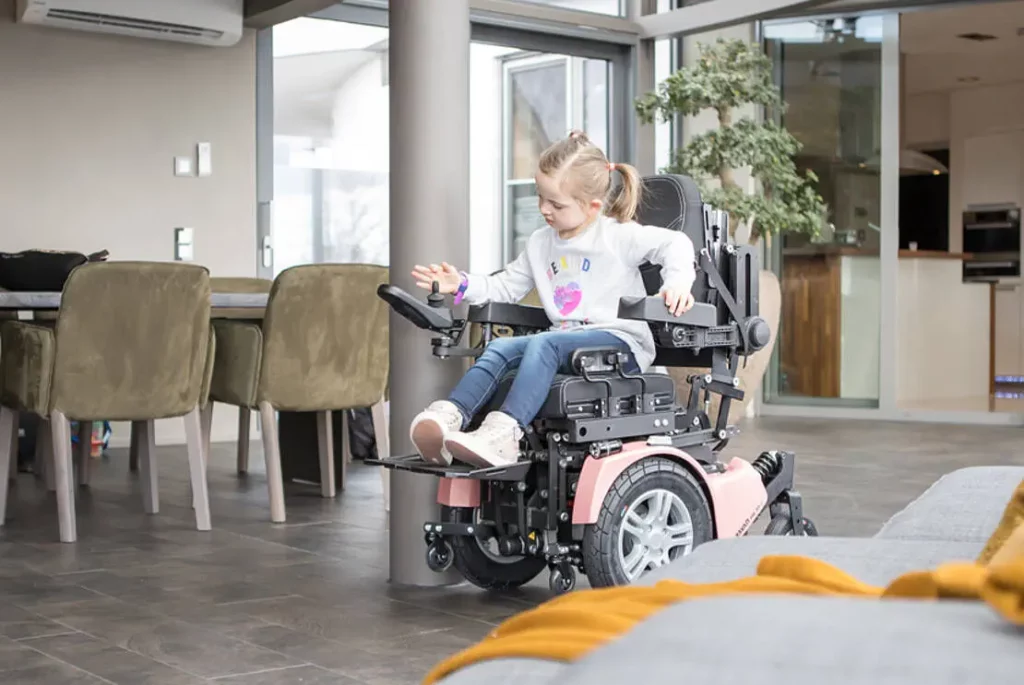 【輪椅王】 A young girl sits in a WCPR 35S Standing Electric Wheelchair indoors, positioned near a dining table and sofa.