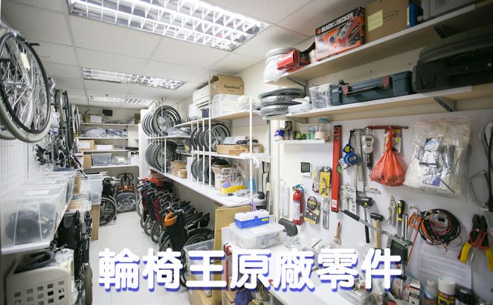 輪椅王 雜亂的車間裡擺滿了各種輪椅零件和工具，包括輪胎、座墊和輪椅維修用的機械部件。架子上擺滿了物品，左側部分可見自行車。